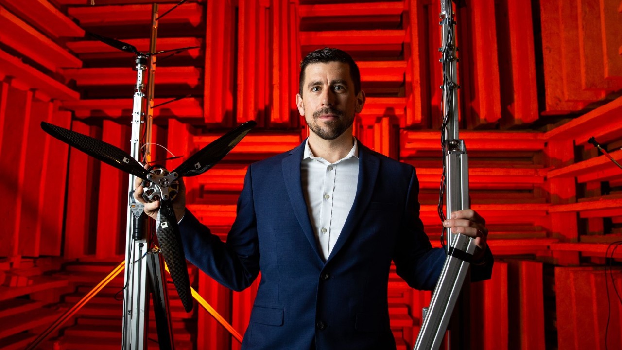 Professor holds propeller pieces and poses for camera in room with red sound-dampening walls behind