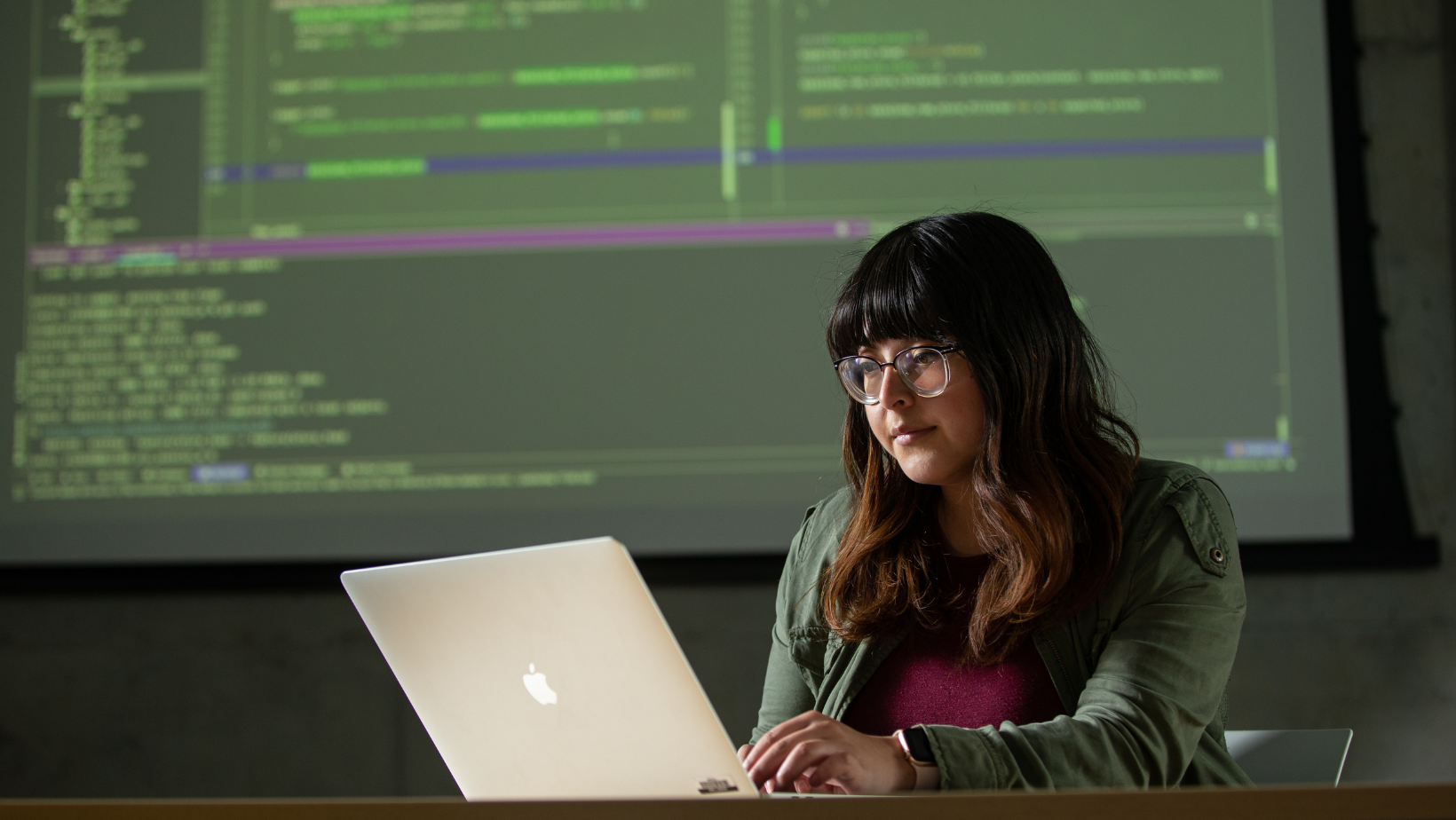 student works at a desk on a laptop with a projection screen of code behind her on the wall