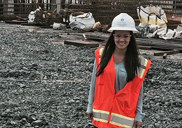 Luana Pego, architectural engineering class of 2017, pictured on a construction site wearing a neon orange safety vest, a hardhat, and safety glasses.