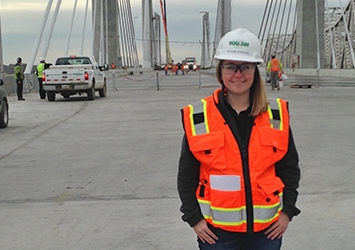 Taylor Bonifant, construction management class of 2018, pictured in a hardhat, neon orange safety vest and safety glasses in front of a bridge construction site.